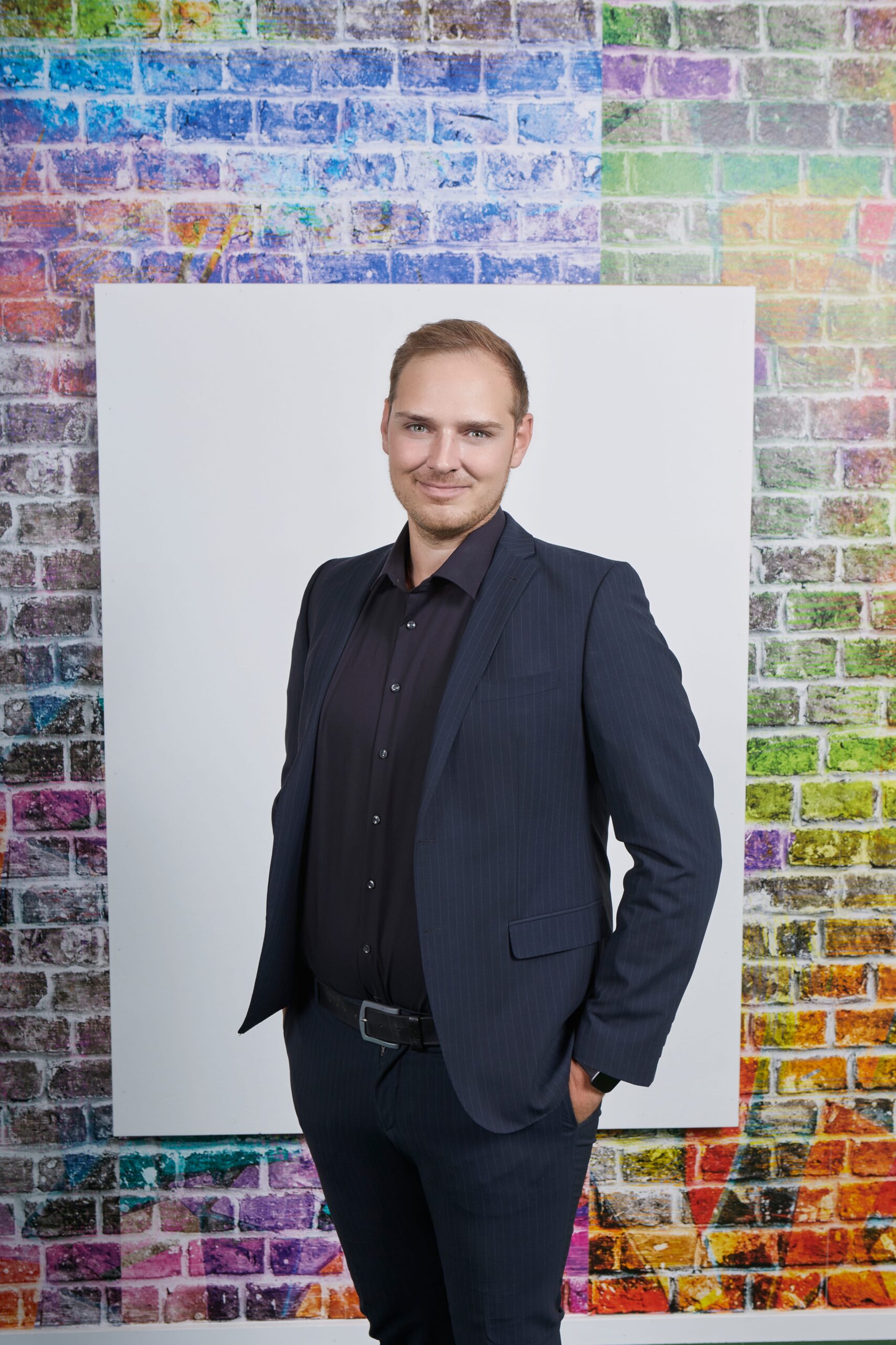 Mann im Anzug steht vor einer bunten Backsteinwand mit einem leeren weißen Rahmen.