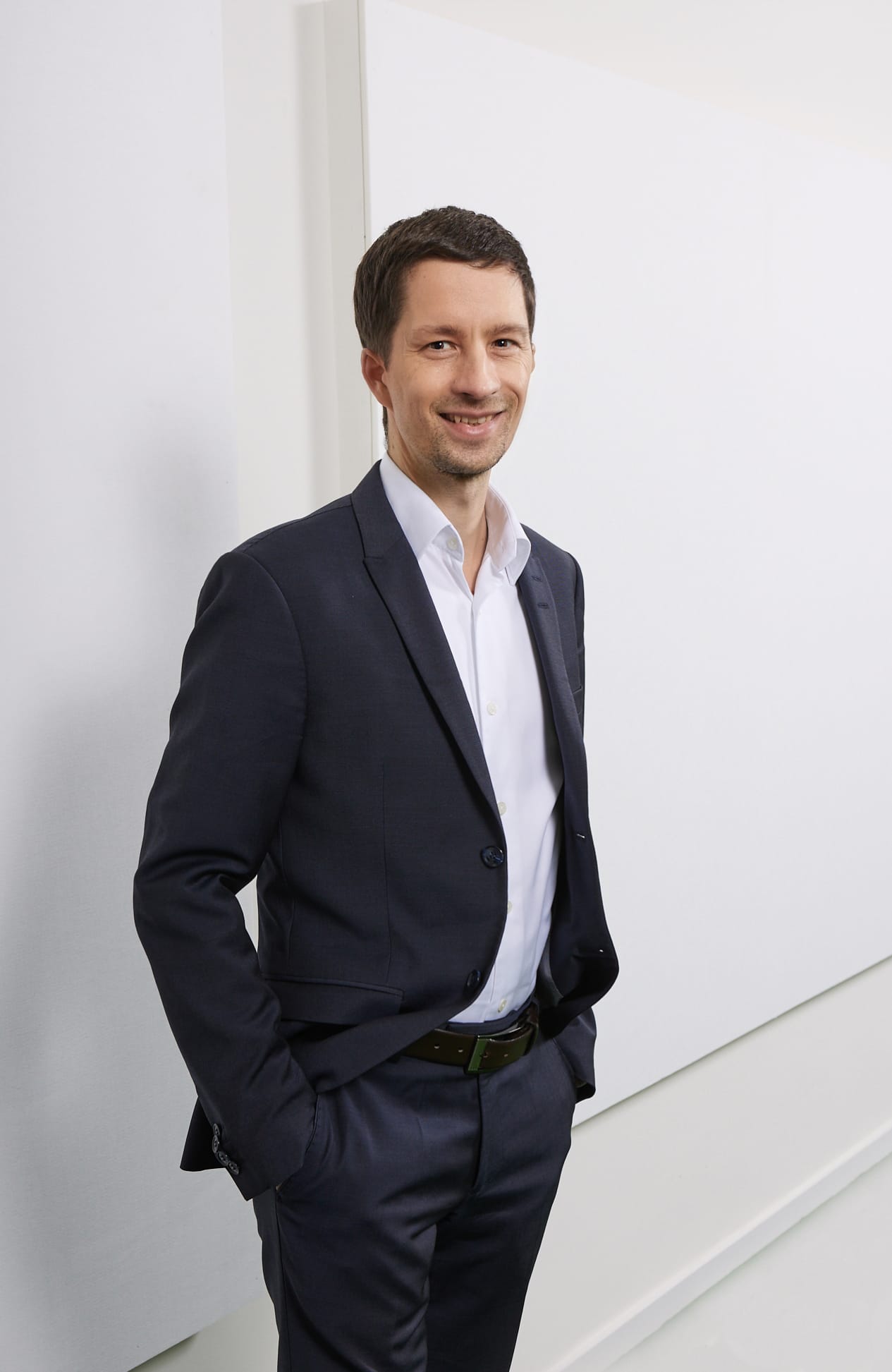 Ein Mann in einem dunklen Anzug lächelt, steht mit einer Hand in der Hosentasche vor einer weißen Wand.