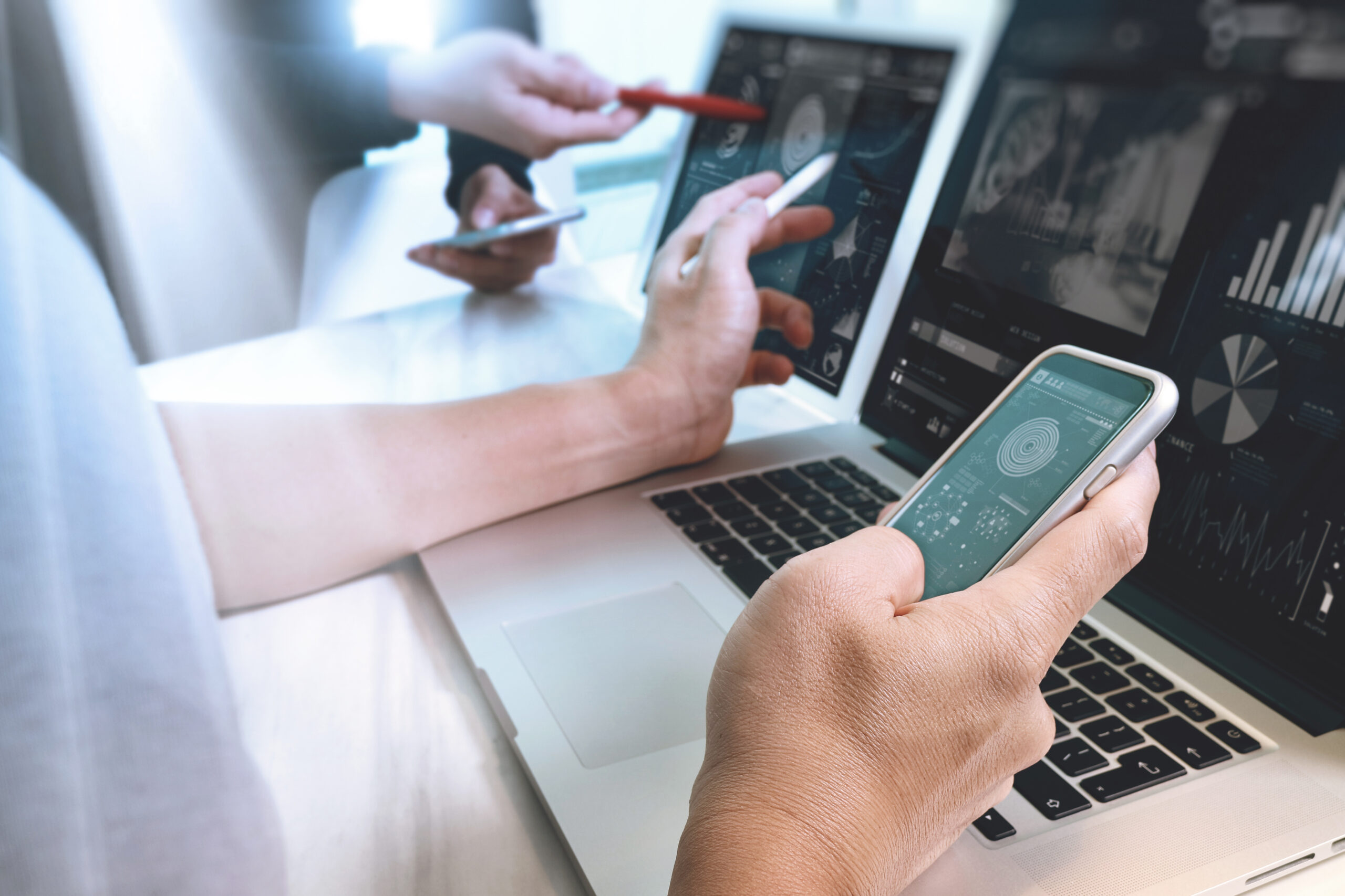 Person h&auml;lt ein Smartphone in der Hand, w&auml;hrend auf einem Laptop Datenanalysen und Diagramme sichtbar sind.