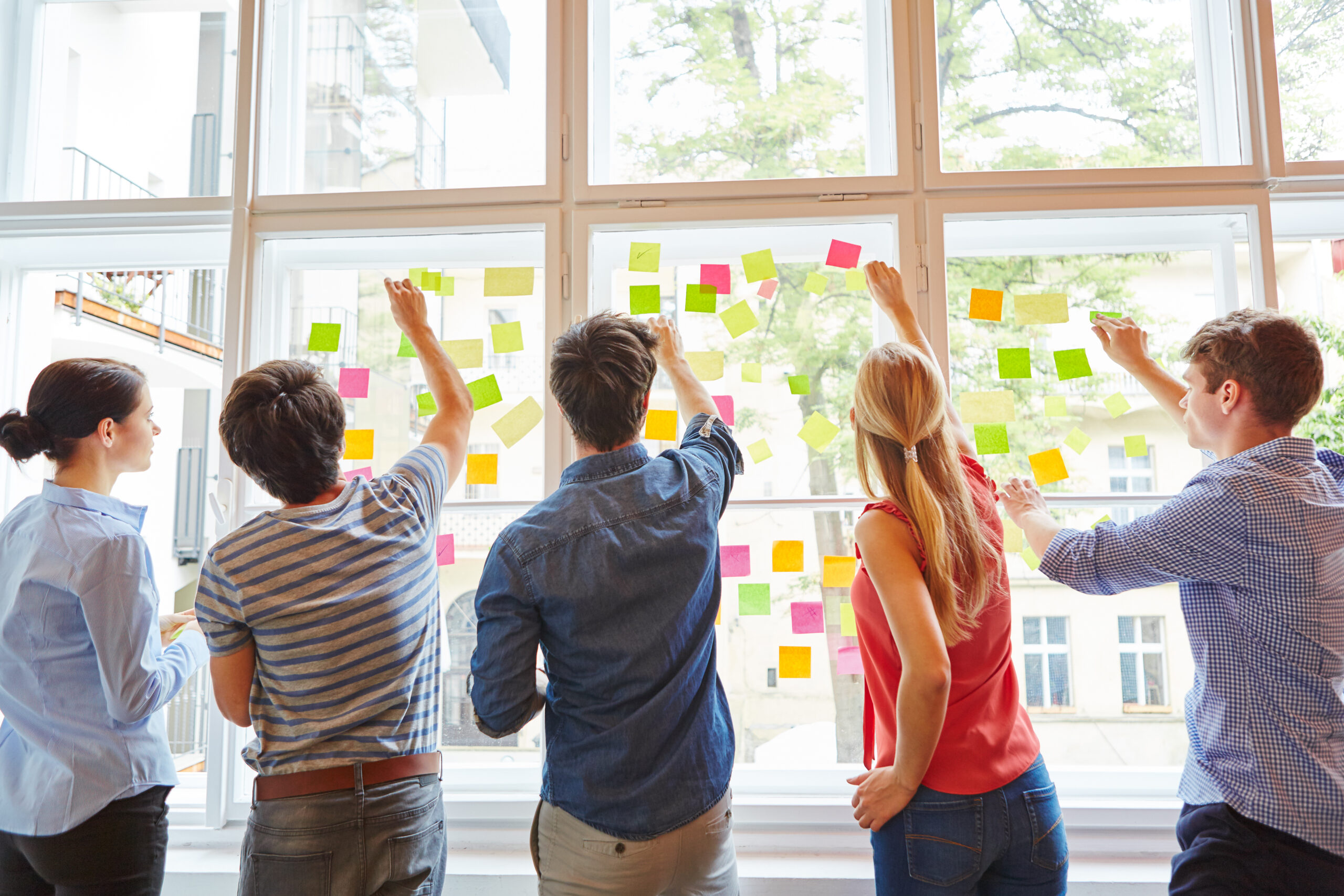 Eine Gruppe von f&uuml;nf Personen klebt bunte Haftnotizen an ein gro&szlig;es Fenster in einem modernen B&uuml;ro.