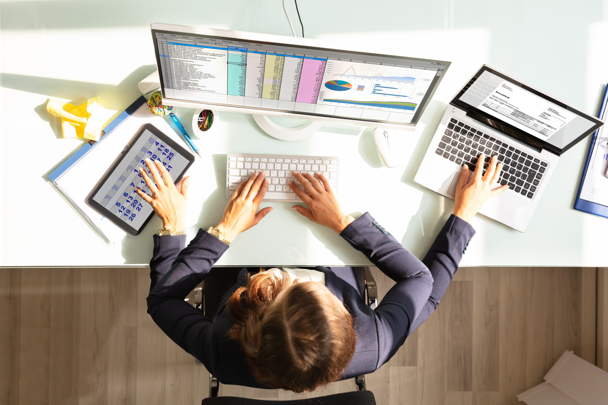 Frau arbeitet an einem Schreibtisch mit zwei Monitoren, Laptop und Tablet, umgeben von Dokumenten und B&uuml;robedarf.